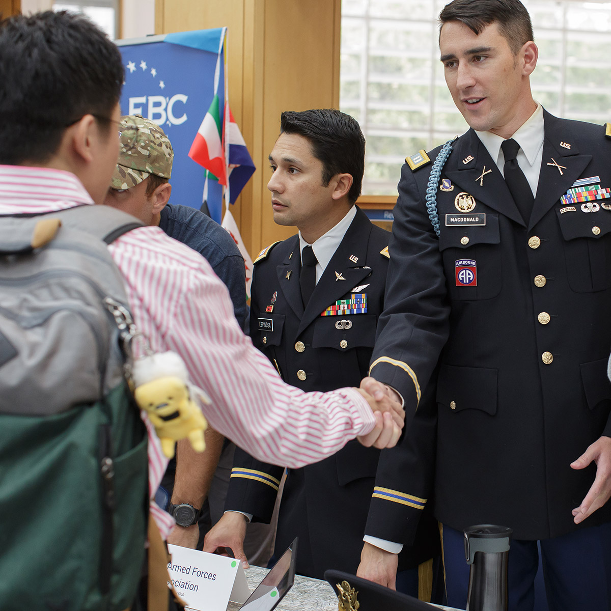 A student wearing a military uniform shakes the hand of another student dressed in civilian clothes. The interaction is happening at a table for the Duke Armed Forces Association at Duke University's Fuqua School of Business
